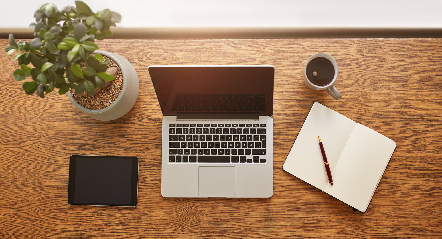 R Facilities - Dicas para deixar o seu ambiente de trabalho organizado Foto por cima de uma mesa de trabalho com um notebook, um caderno, caneca com café e uma vaso de plantas