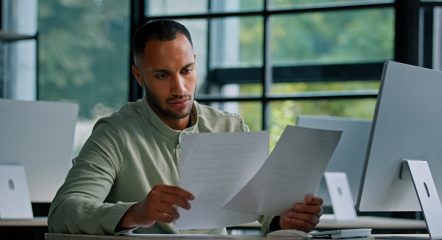 R Facilities - Para quem é a terceirização Homem sentado na frente computador enquanto segura folhas de papel.