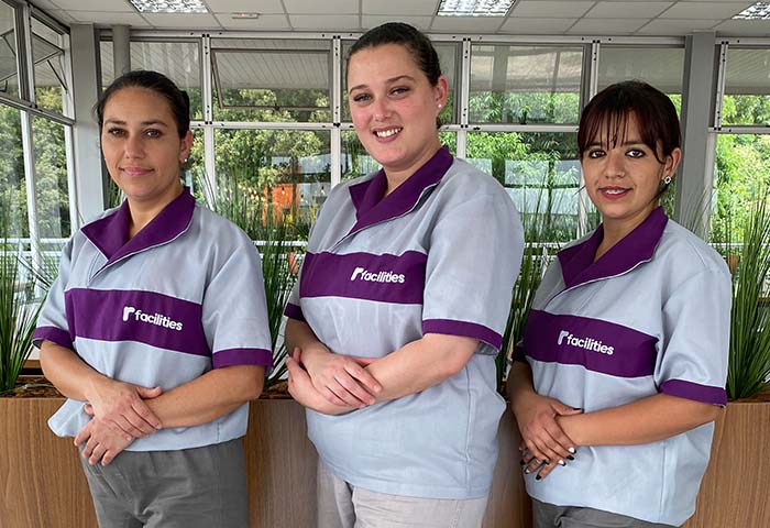 Três colaboradoras uniformizadas da R Facilities, sorrindo e posicionadas em um ambiente corporativo, representando padronização, profissionalismo e cuidado com a limpeza.